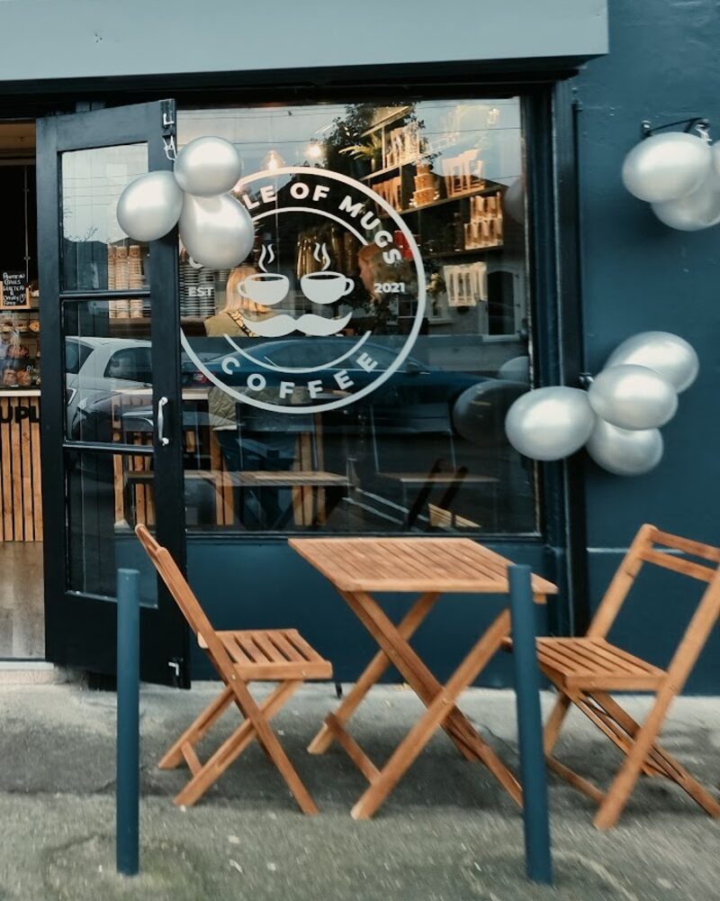 Wooden folding tables and chairs on the pavement outside the Couple Of Mugs Coffee window.