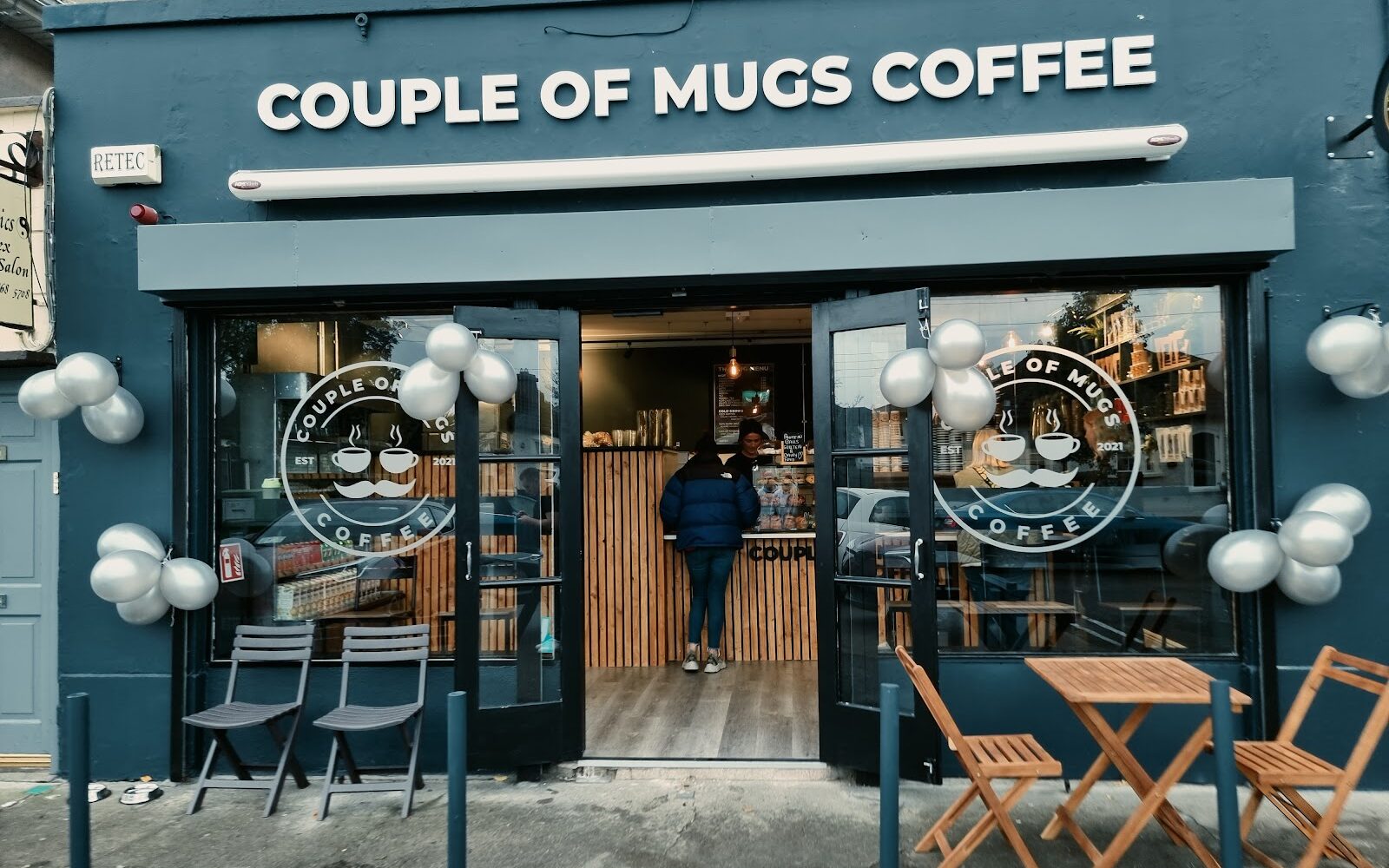 The navy-blue Couple Of Mugs Coffee shopfront on Nephin Road with open doors, a timber-slatted counter inside and tables on the pavement.